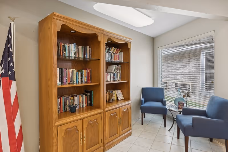 A small reading nook with two blue armchairs and a round side table holding decorative items, positioned next to a large window with blinds. To the left, there is a wooden bookshelf filled with books and an American flag standing in the corner.