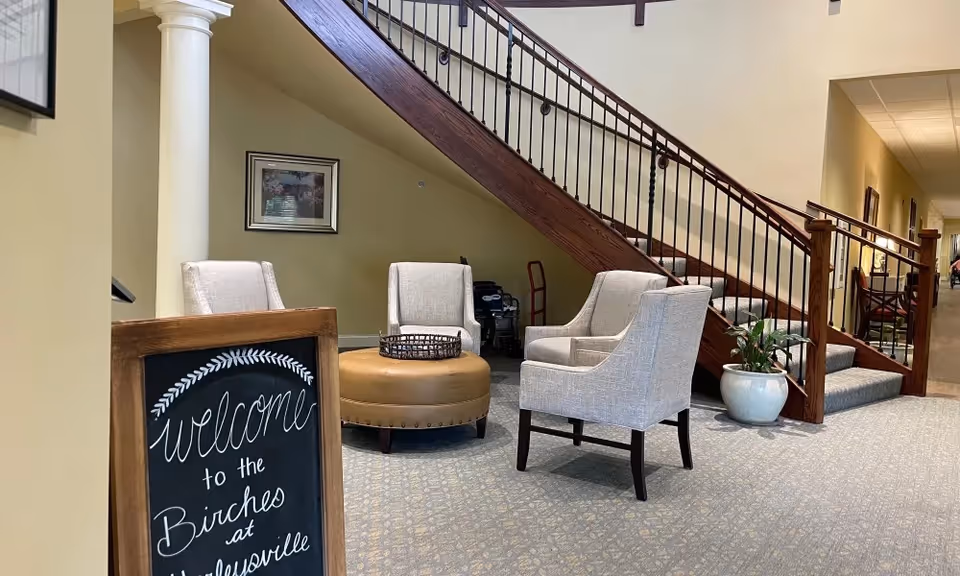 A welcoming lounge area with upholstered armchairs and a round ottoman under a wooden staircase and a chalkboard sign reading welcome to the Birches at Harleysville.
