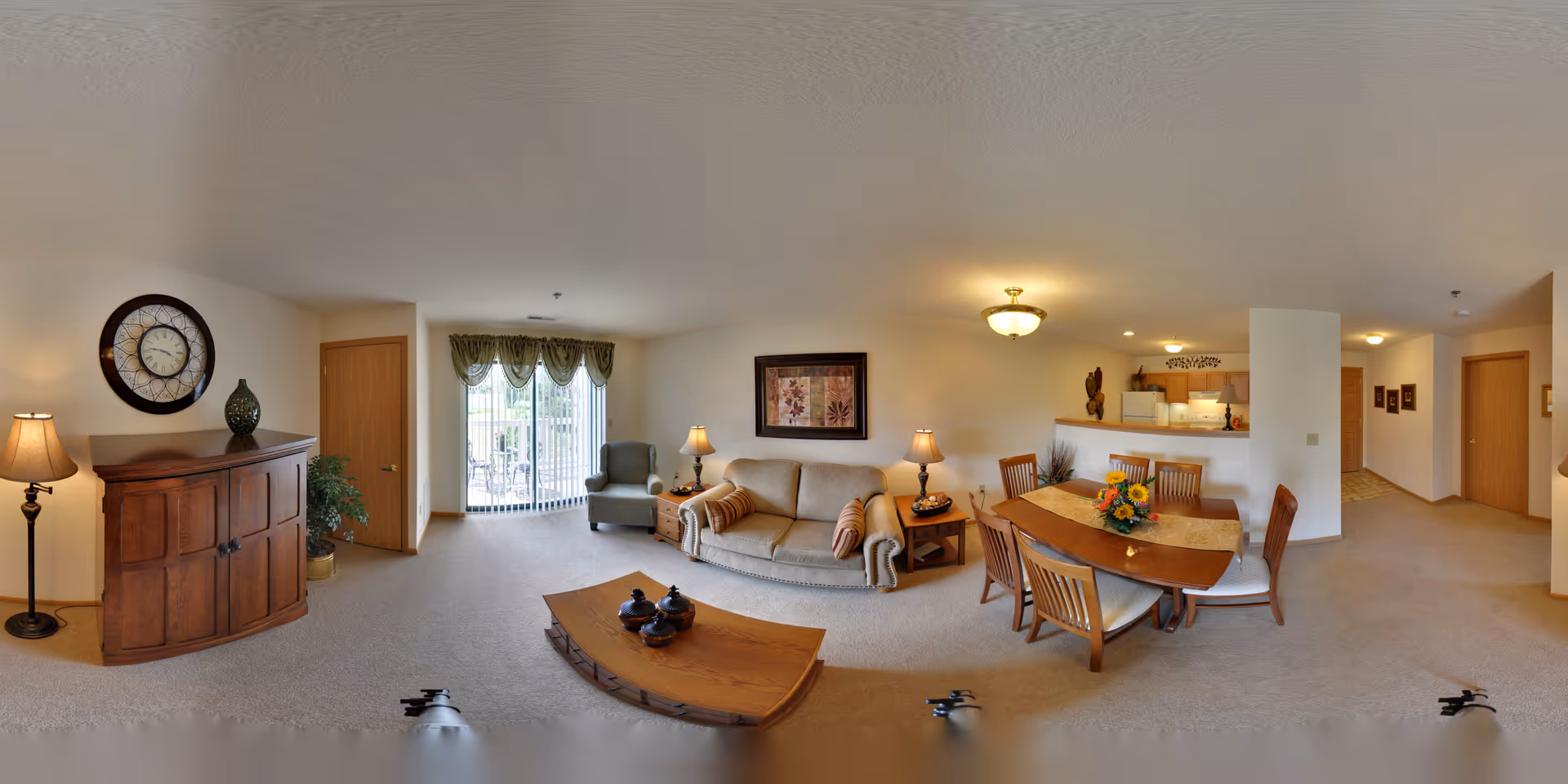 A spacious senior living facility common area featuring a beige sofa with two striped pillows, a wooden coffee table with decorative items, a wooden dining table with six chairs and a floral centerpiece, a wooden cabinet with a large clock and vase on top, a floor lamp, an armchair near sliding glass doors with green valances leading to an outdoor patio, and a kitchen area with wooden cabinets and a pass-through counter.