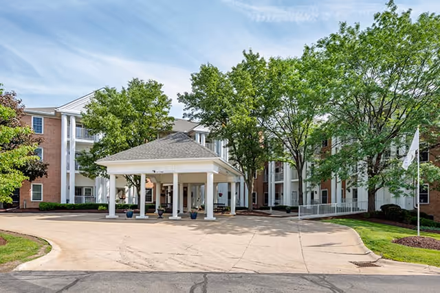 Front entrance of a brick senior living facility featuring a covered porte-cochere, circular driveway, and trees.