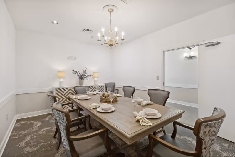 A well-lit dining room with a rectangular wooden table set for six people. The table has plates, bowls, glasses, and napkins neatly arranged. Surrounding the table are six upholstered chairs with wooden arms. In the background, there is a sideboard with two lamps and a decorative vase with branches. A chandelier hangs from the ceiling, and the walls are painted white with a beige lower section and white trim. An open door leads to another room with similar decor.