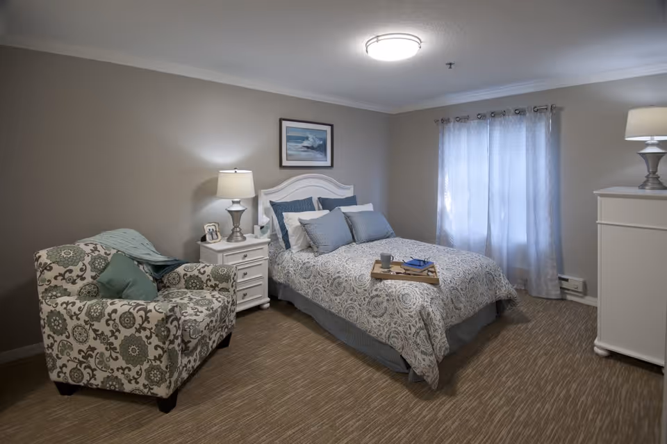 A cozy bedroom with a patterned armchair on the left, a white nightstand with a lamp and picture frame, a bed with multiple pillows and a patterned bedspread, a tray with a cup and book on the bed, sheer curtains covering a window, and a white dresser with a lamp on the right.