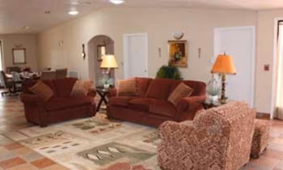 A furnished common living area with red sofas, a patterned armchair, side tables with lamps, and a large rug.