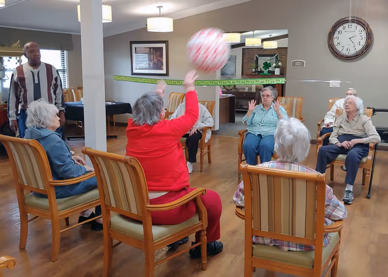 A group of elderly people sitting in a circle of wooden chairs with striped cushions in a common room. One person in a red outfit is raising their arms to catch or throw a large ball. A man stands nearby observing the activity. The room has wooden flooring, a large clock on the wall, and framed pictures.