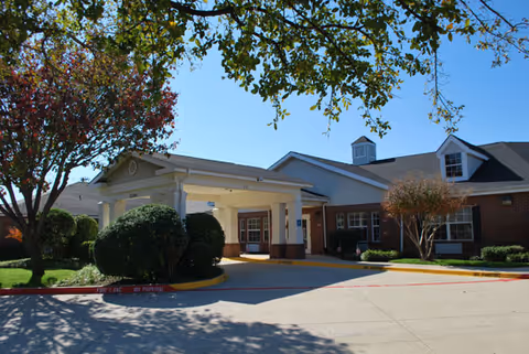 Exterior view of Brookdale Farmers Branch senior living facility showing a single-story brick building with a covered entrance, surrounded by trees and bushes under a clear blue sky.