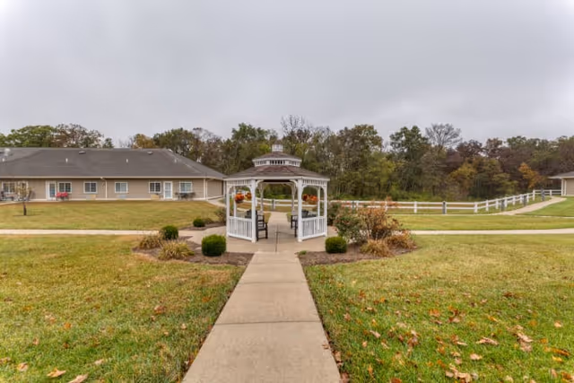 A white wooden gazebo with hanging flower baskets is situated at the center of a paved walkway in a grassy outdoor area. Surrounding the gazebo are bushes and landscaped plants. In the background, there are single-story beige buildings with windows and a white fence, with trees further behind under a cloudy sky.