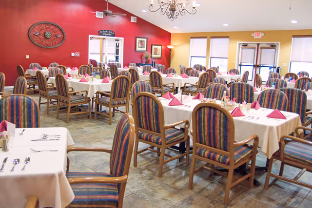 Spacious dining room with multiple tables set with white tablecloths and pink napkins and striped upholstered chairs against a red accent wall.