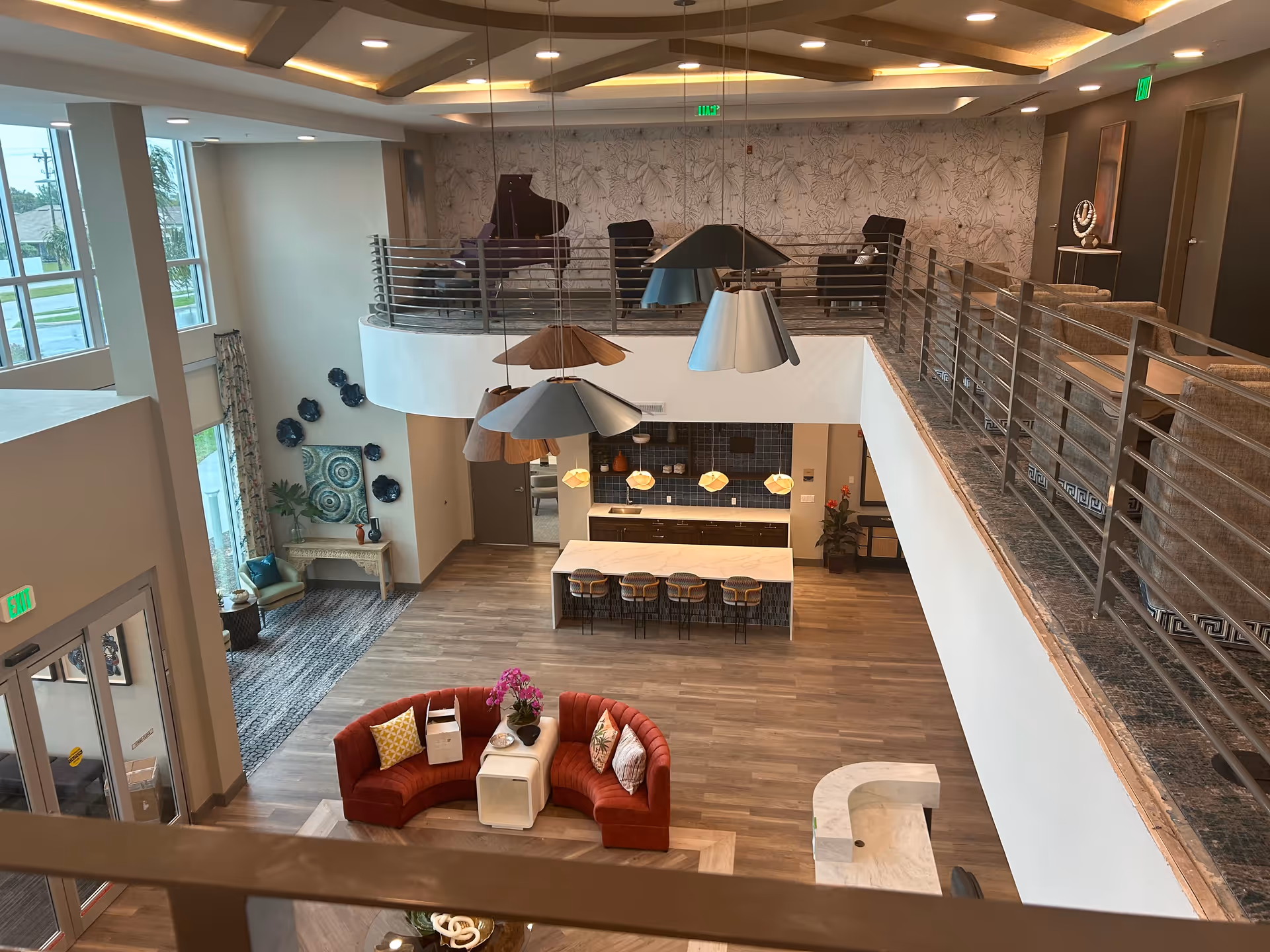 View from an upper level overlooking a spacious, modern common area in a senior living facility. The area features a curved red sofa with pillows around a small white table, a kitchen island with bar stools, pendant lights, and a mezzanine with seating and a grand piano. Large windows allow natural light to fill the space, and decorative wall art and plants add to the welcoming atmosphere.