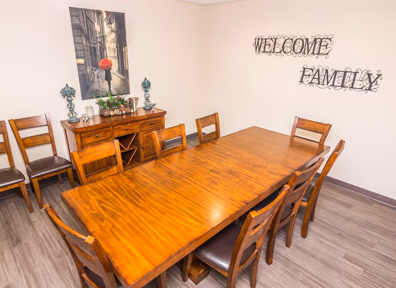 A dining room with a long wooden table surrounded by eight wooden chairs with dark cushions. Against the wall is a wooden sideboard with decorative items and a framed picture above it showing a person holding a red umbrella walking down a street. On the wall, there is a decorative metal sign that reads 'WELCOME FAMILY'. The floor is covered with light-colored wood laminate.