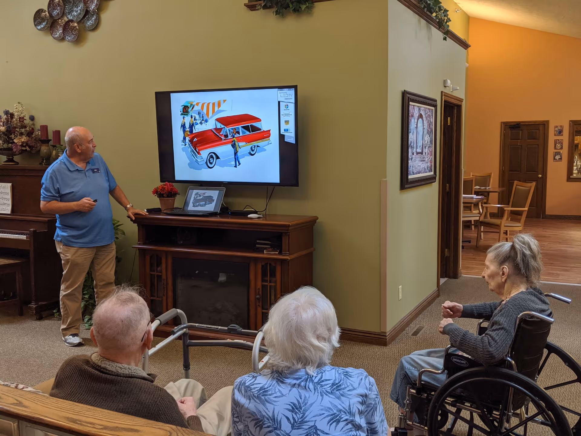 A man in a blue polo shirt stands next to a TV mounted on a green wall, giving a presentation about a red vintage car to three elderly people seated in front of him. One elderly woman is in a wheelchair, and the room has warm lighting with wooden furniture and framed pictures on the walls.