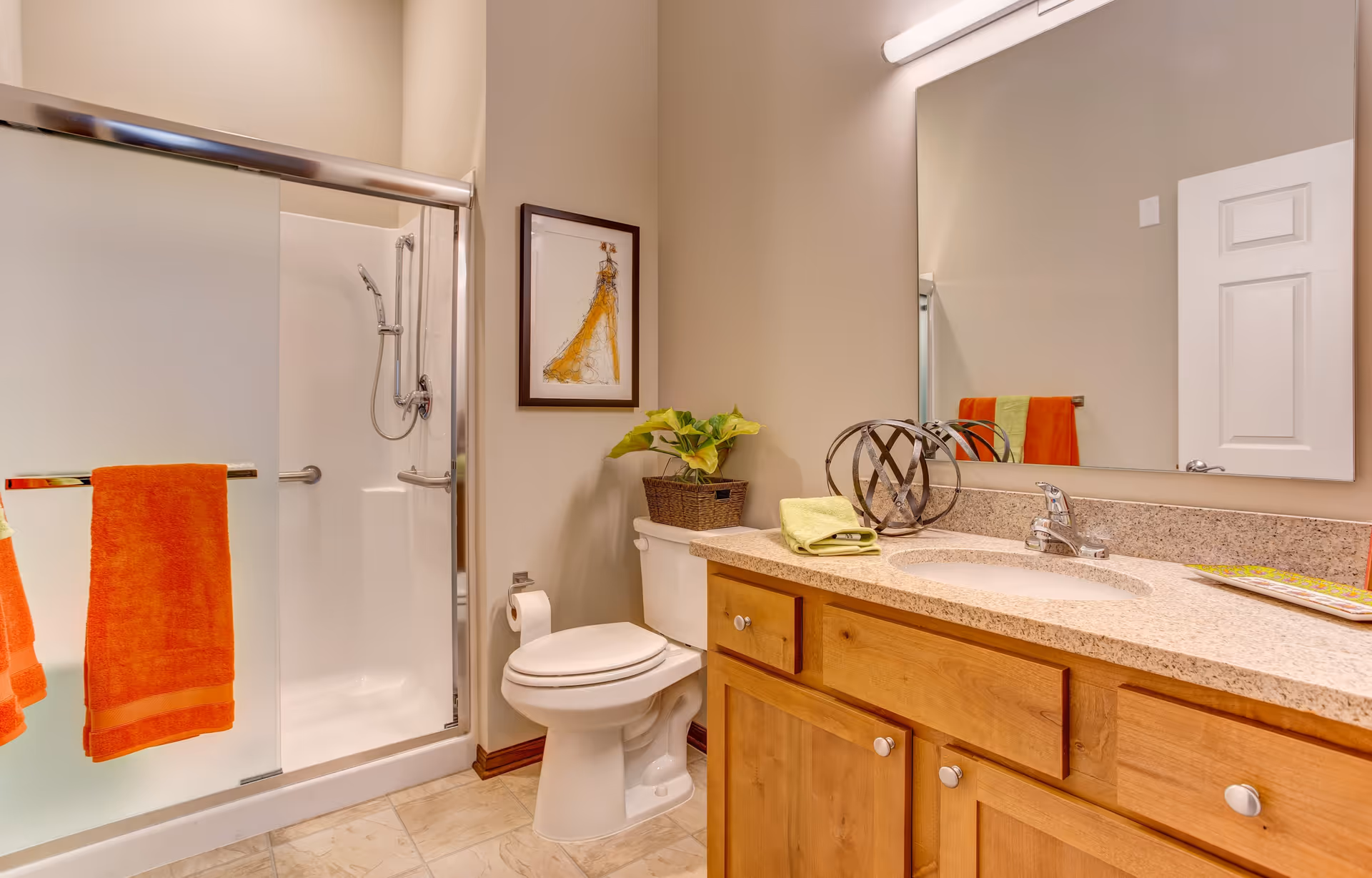 A clean and well-lit bathroom featuring a glass-enclosed shower with a silver frame and a mounted showerhead. There are two orange towels hanging on the shower door handle. Next to the shower is a white toilet with a roll of toilet paper on the wall. Above the toilet is a framed artwork of a dress. A wooden vanity with a granite countertop holds a sink with a silver faucet, a green folded towel, a decorative metal orb, a potted plant, and some magazines. A large mirror and a white door are visible in the background.