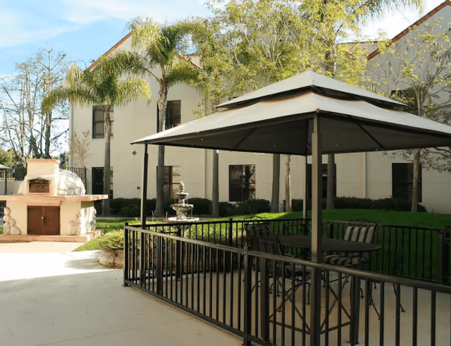Outdoor patio area with a gazebo covering a table and chairs, surrounded by a black metal fence. In the background, there is a two-story building with windows, palm trees, a stone barbecue grill, and a decorative water fountain.