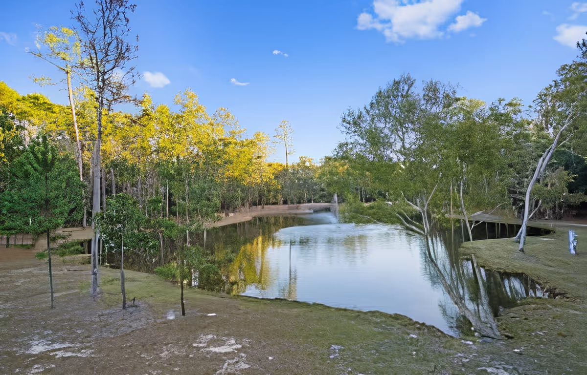 A serene outdoor scene featuring a calm pond surrounded by trees with green and yellow foliage under a clear blue sky.