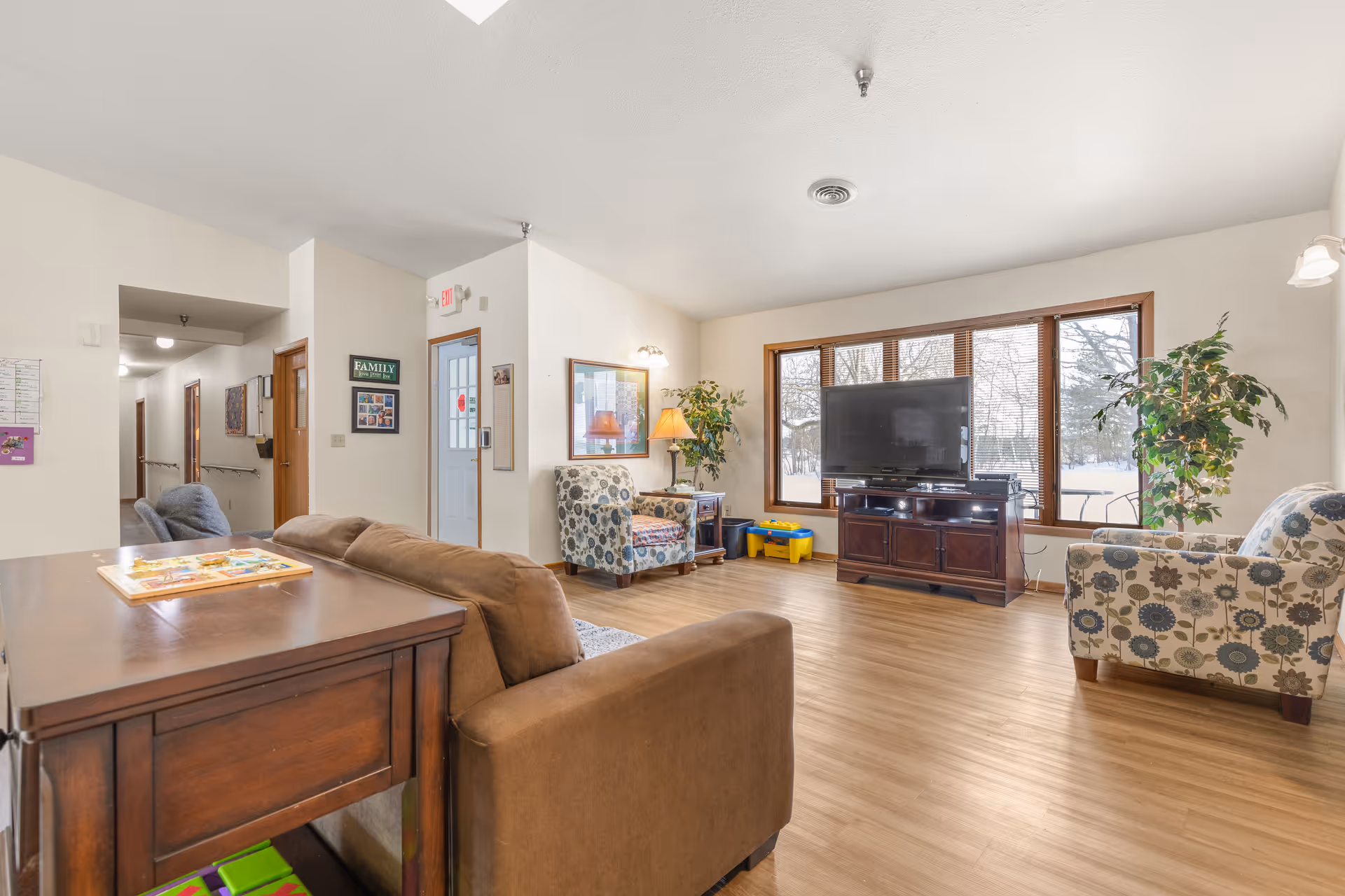 A bright and spacious living room with large windows letting in natural light. The room features a brown sofa, two patterned armchairs, a wooden TV stand with a flat-screen television, a side table with a lamp, and some indoor plants. The floor is wooden, and there is a hallway visible leading to other rooms.