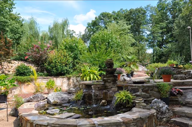 Stone terraced garden with a small pond and waterfall surrounded by potted plants, rock steps, and lush trees.