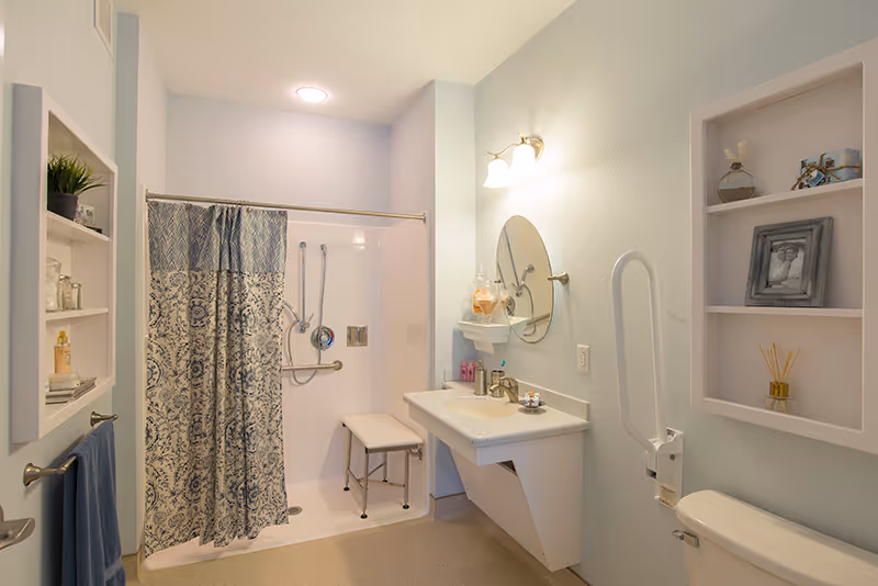 A clean and accessible bathroom with a walk-in shower featuring a patterned shower curtain, a shower bench, and grab bars. There is a wall-mounted sink with a round mirror above it, a light fixture, and shelves with decorative items. A towel hangs on a rack next to the shower.