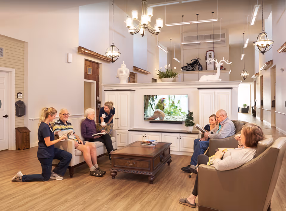 A bright, warmly furnished communal living room with residents and caregivers seated around a coffee table watching a TV and conversing.