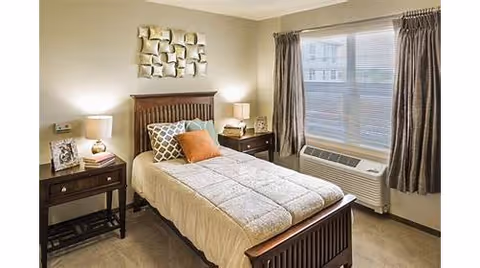 A cozy bedroom with a single wooden bed featuring a beige comforter and decorative pillows. There are two dark wooden nightstands on either side of the bed, each with a lamp and decorative items. A large window with closed blinds and dark curtains is on the right wall, and a modern air conditioning unit is installed below the window. The walls are painted a light beige color, and a decorative wall art piece hangs above the bed.