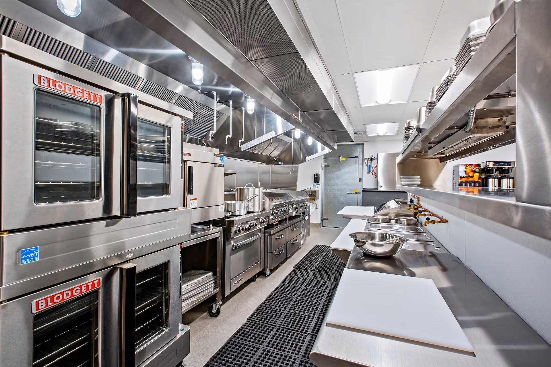 A modern commercial kitchen with stainless steel appliances including Blodgett ovens, stovetops, and preparation counters with cutting boards and mixing bowls. The kitchen is well-lit with overhead lights and has a clean, organized appearance.