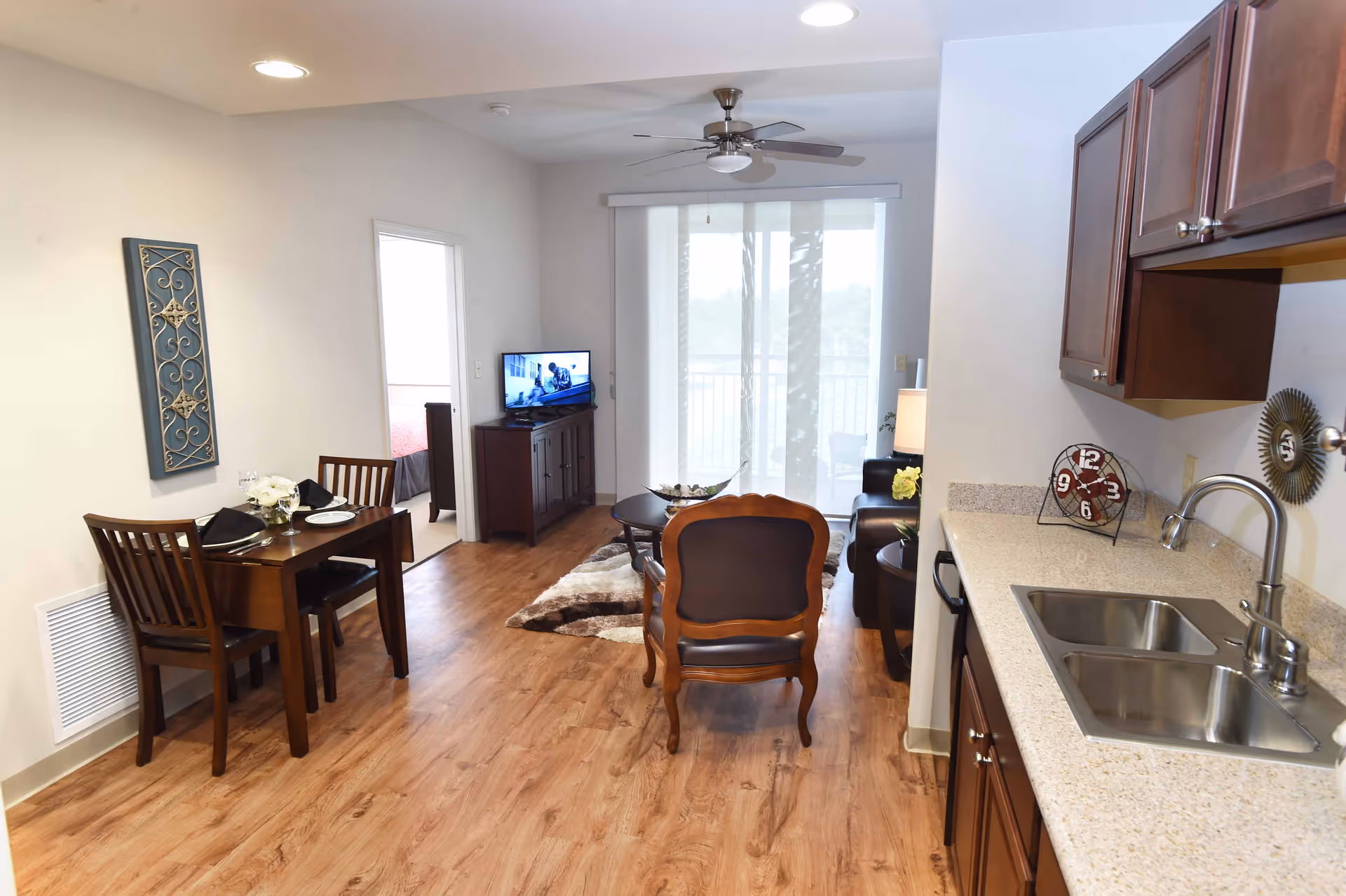 Interior view of a senior living facility apartment showing a small dining area with a wooden table and two chairs set with plates and napkins, a living room with a wooden armchair, a coffee table, a TV on a stand, and sliding glass doors leading to a balcony. The kitchen area with a double sink and wooden cabinets is visible on the right side.