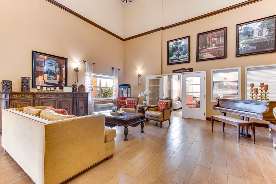 A spacious senior living facility common area with high ceilings, beige walls, and wooden flooring. The room features a beige sofa with red cushions, two armchairs with red patterned cushions, a wooden coffee table, and a wooden sideboard with decorative items. There is a popcorn machine near the window, and a grand piano with a bench on the right side. Above the double doors labeled 'Fireside Room' are three framed pictures of outdoor fountains and gardens. The room is well-lit with natural light coming through the windows and wall sconces.