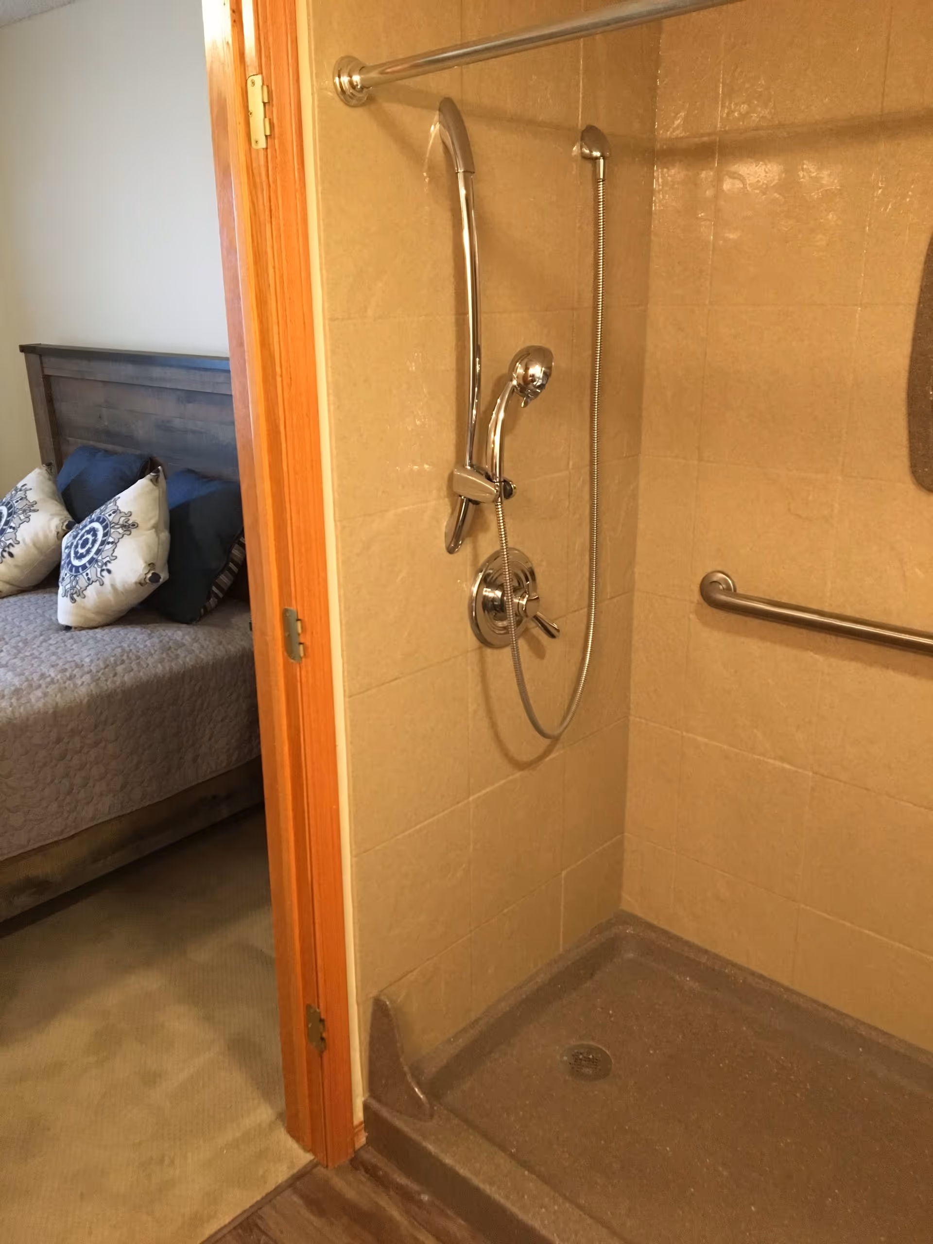 View of a beige tiled shower with a chrome handheld showerhead and grab bar, adjacent to a bedroom with a wooden bed frame and decorative pillows.