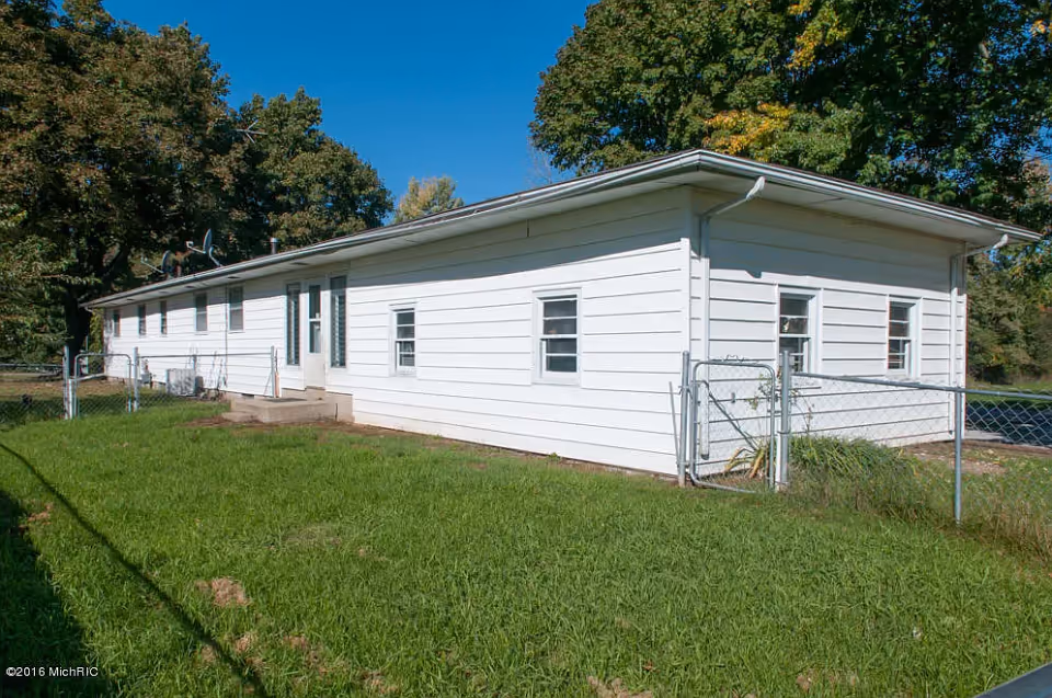 Single-story white building with multiple windows and a small fenced yard, surrounded by green grass and trees under a clear blue sky.