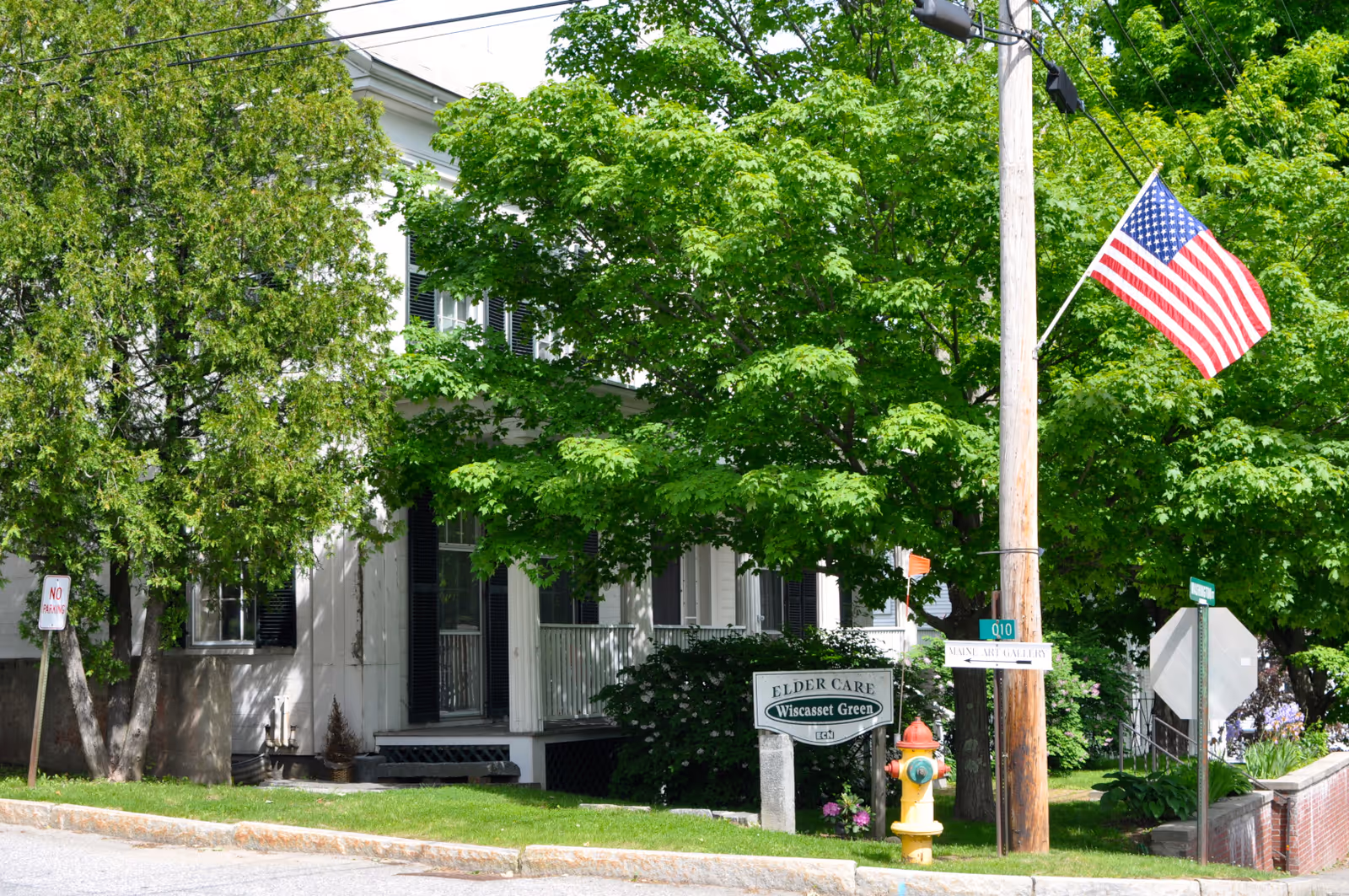 Exterior view of a white elder care facility partially obscured by green leafy trees. There is a porch with black shutters on the windows. A yellow fire hydrant and an American flag on a wooden utility pole are visible in the foreground. A sign near the building reads 'Elder Care Wiscasset Green'.