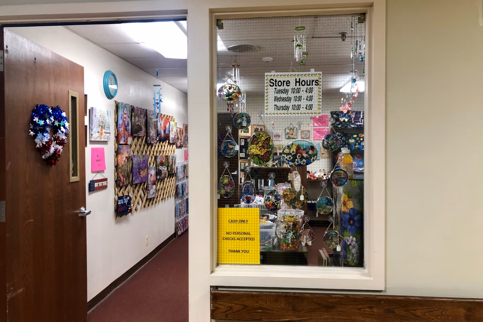 Interior hallway view of Abbott Terrace Health Center showing a wooden door decorated with a red, white, and blue star wreath on the left. The hallway walls are adorned with various colorful artworks and crafts. A window on the right reveals a small store area with hanging decorative items and signs displaying store hours and payment policies.