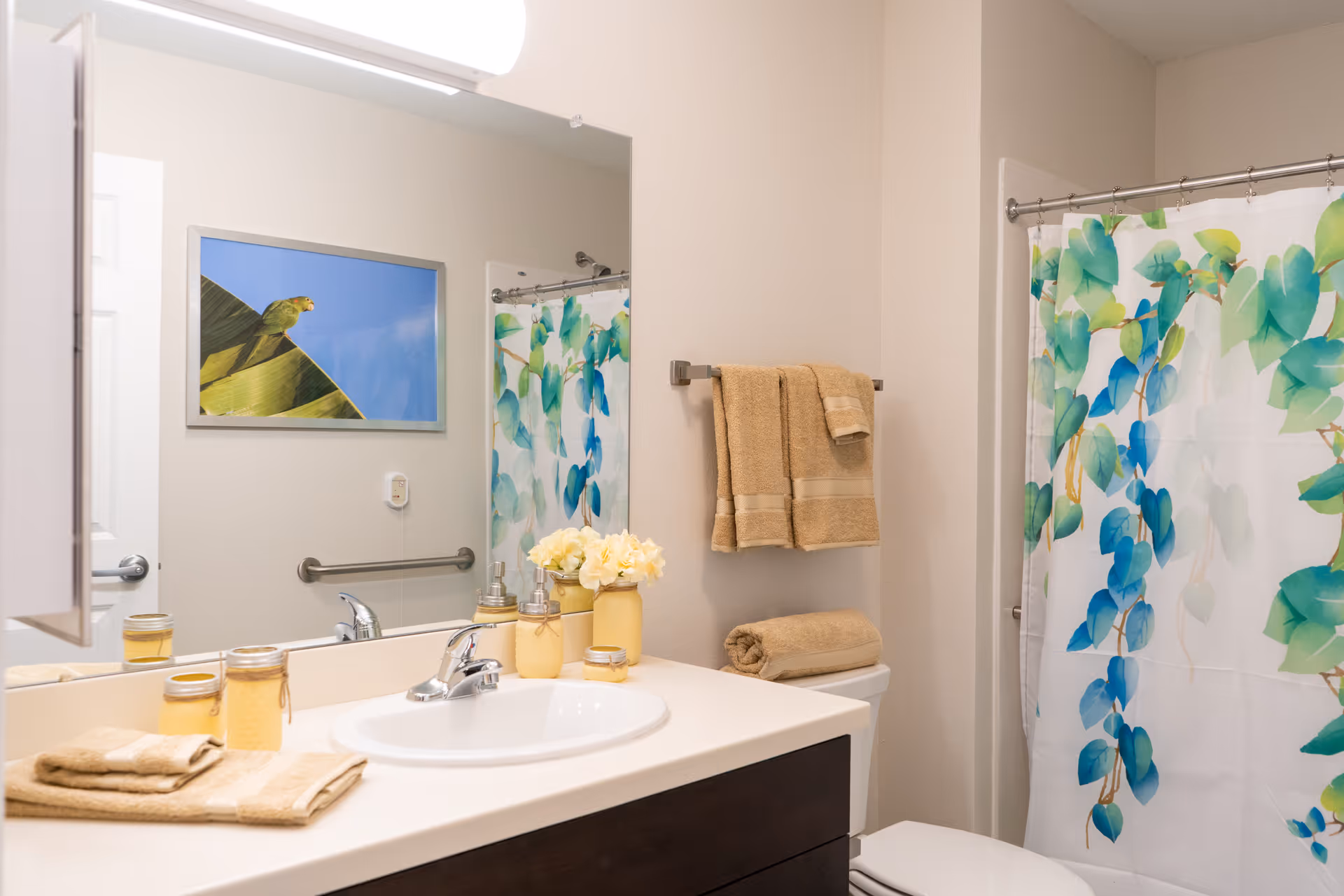 A clean and bright bathroom featuring a white sink with a silver faucet set in a dark wood vanity. Beige towels are neatly folded and hung on a towel rack above the toilet. A shower curtain with a green and blue leafy pattern hangs on a silver rod. A framed picture of a green parrot on a leaf against a blue sky is reflected in the mirror above the sink. Small decorative jars and a vase with white flowers are placed on the countertop.