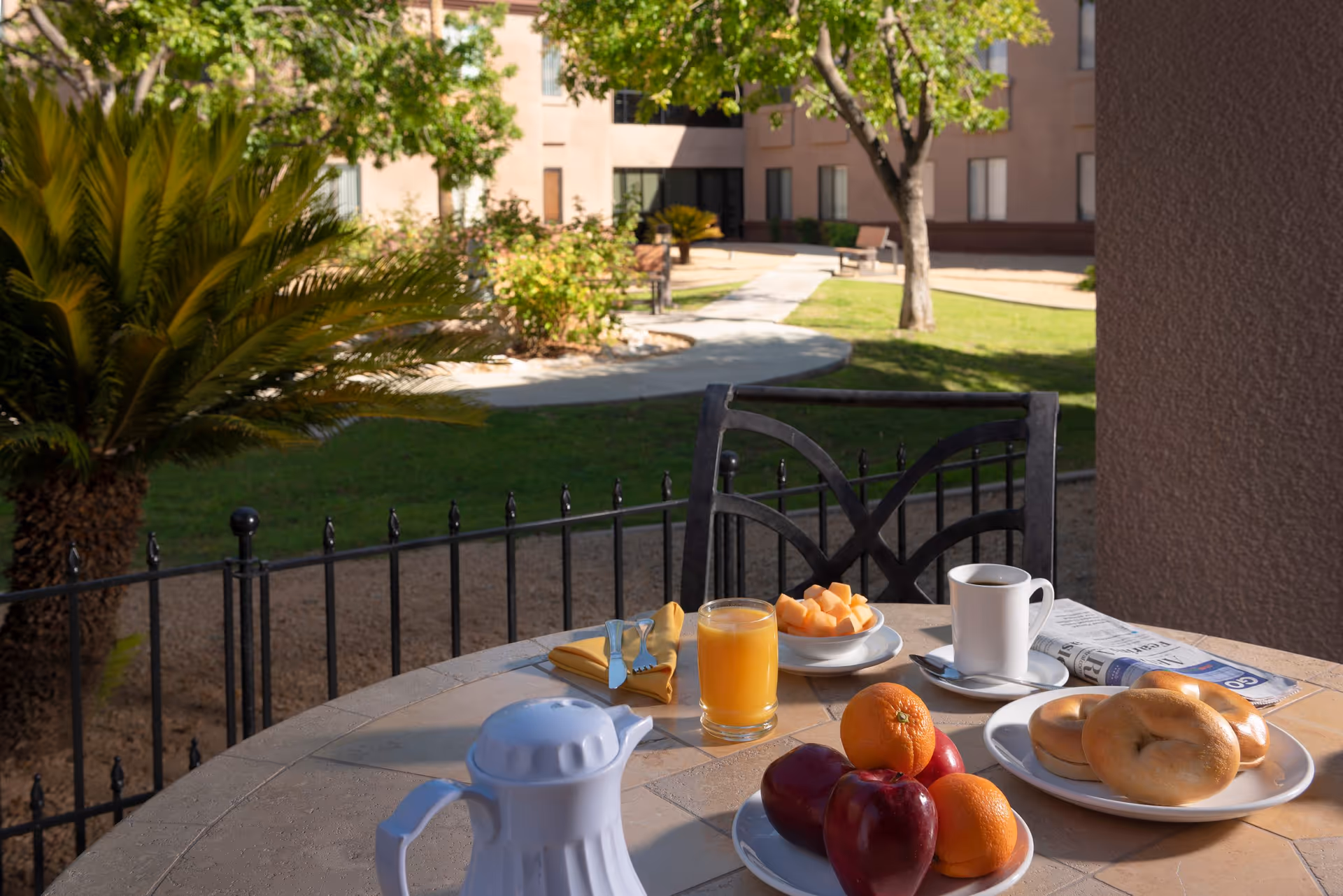 Outdoor patio table set with breakfast items including a white coffee pot, a glass of orange juice, a plate of cantaloupe slices, a cup of coffee, a newspaper, a plate with two bagels, and a plate with apples and oranges. The table is on a patio overlooking a garden area with trees, bushes, and a pathway leading to a building in the background.