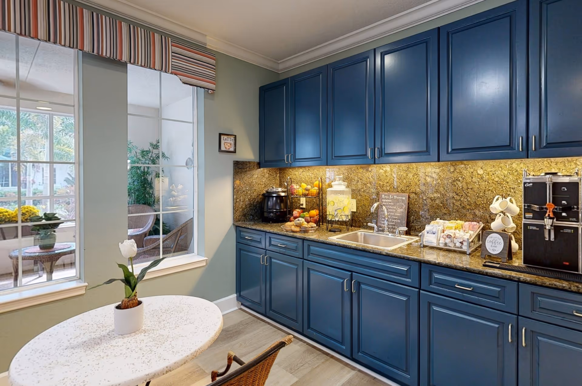 A cozy kitchenette area with blue cabinets and granite countertops. There is a small round table with a white flower in a pot in the foreground. The countertop features a sink, a coffee machine, a water dispenser, a fruit basket, and various coffee supplies. Large windows with striped valances look out to an outdoor patio area with wicker chairs and plants.