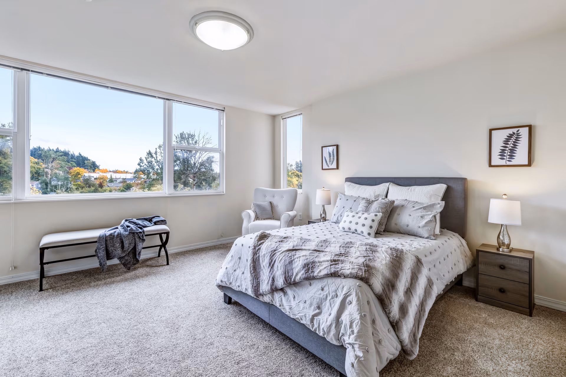 Bright bedroom with a large window showing a view of trees outside. The room features a neatly made bed with multiple pillows and a gray upholstered headboard, a white armchair with a striped pillow, two framed leaf prints on the wall, a wooden nightstand with a lamp, and a bench with a folded blanket near the window. The floor is carpeted in a light beige color.