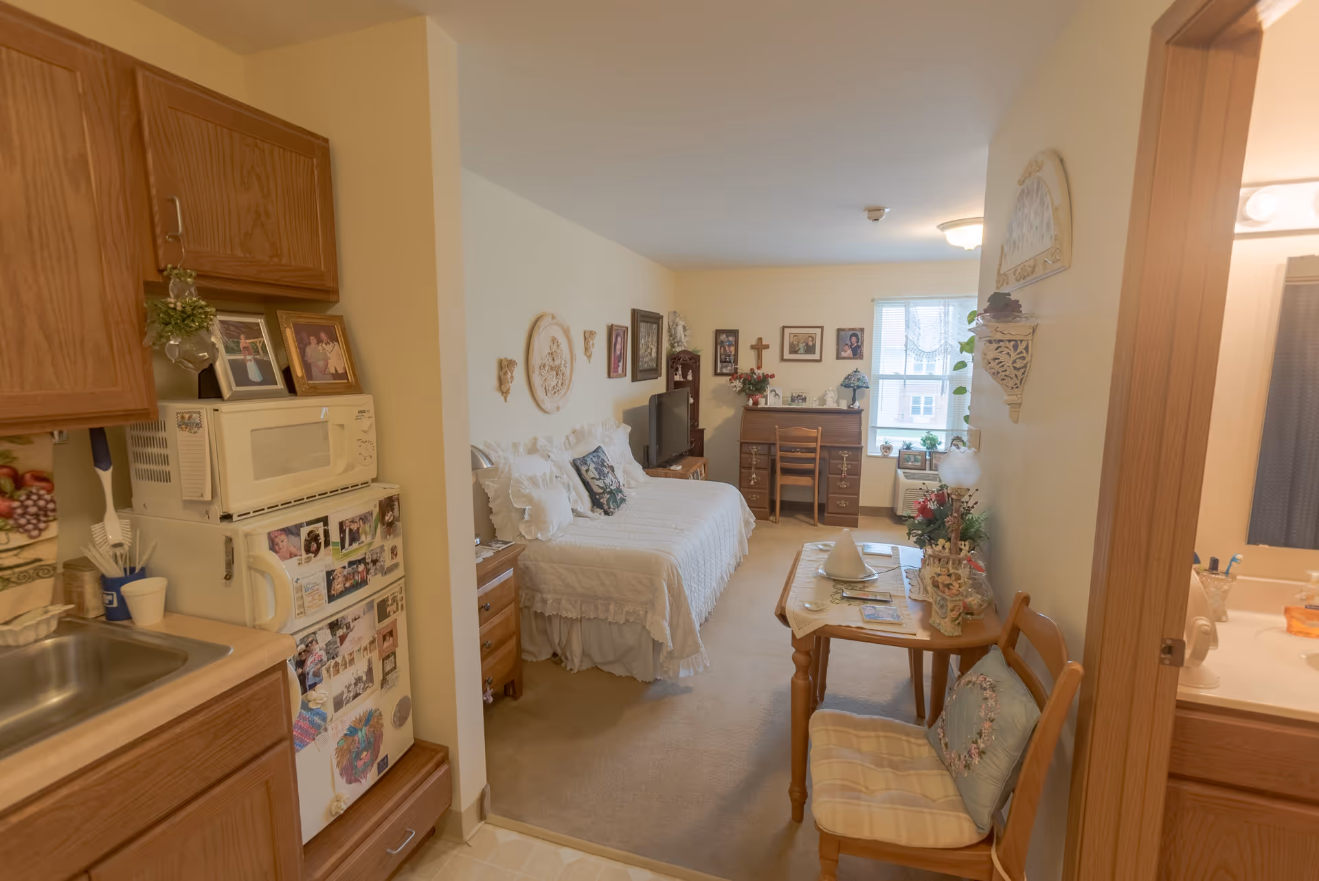 A cozy studio apartment in a senior living facility featuring a small kitchenette with a sink, microwave, and refrigerator adorned with photos and magnets. The living and sleeping area includes a daybed with white ruffled bedding, a wooden desk with a chair near a window, a TV on a stand, and various decorative wall hangings and framed photos. A small wooden dining table with two chairs is positioned near the entrance to a bathroom, which has a sink and mirror visible.