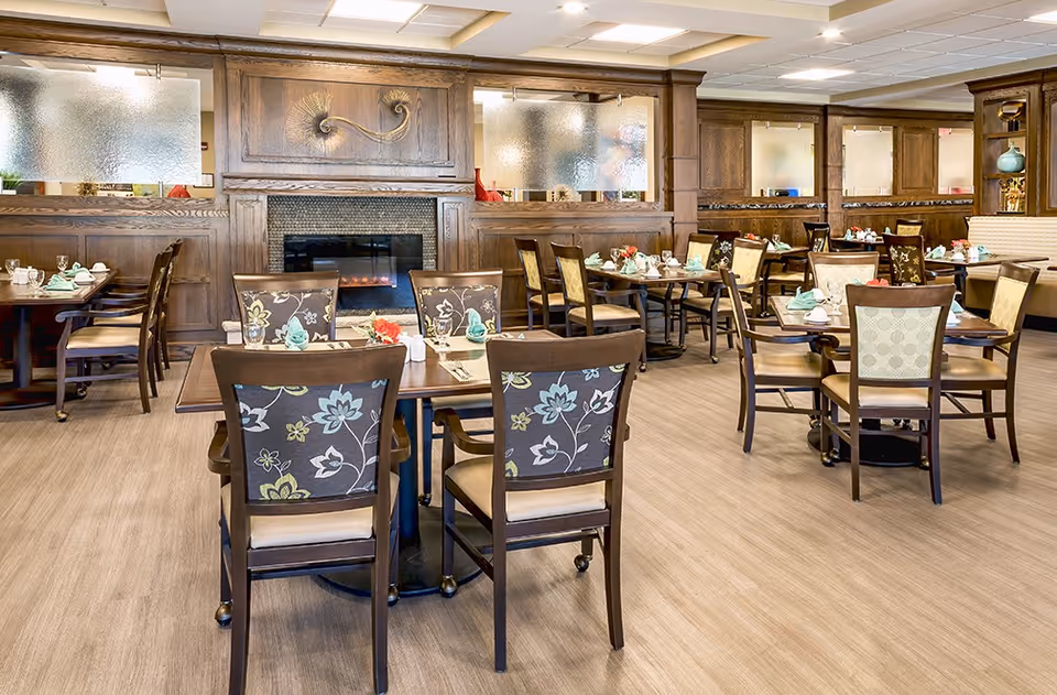 A spacious dining room with multiple wooden tables and chairs arranged neatly. Each table is set with napkins, glasses, and small flower arrangements. The room features a wooden paneled wall with frosted glass windows and a decorative fireplace in the center. The floor has a light wood finish, and the overall ambiance is warm and inviting.