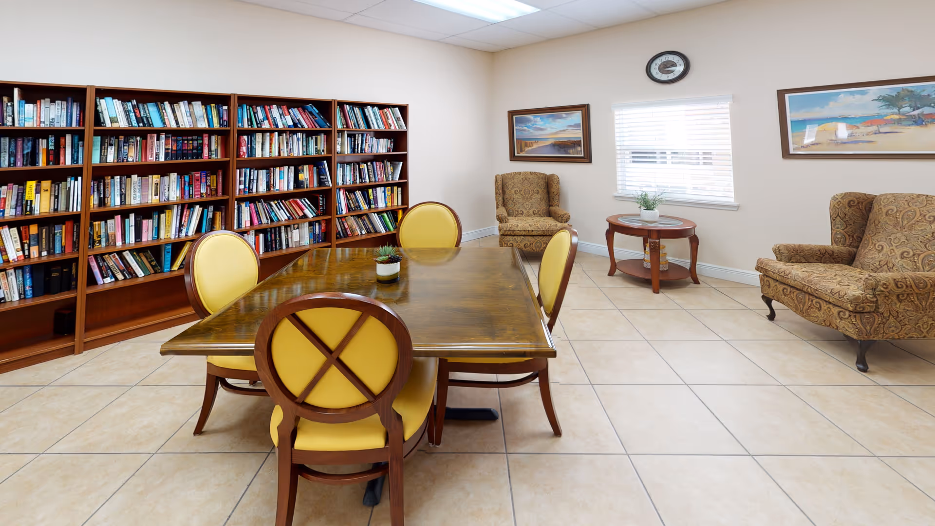 Communal reading room with a wooden table surrounded by yellow-cushioned chairs, bookshelves along the left wall and upholstered armchairs by a window.