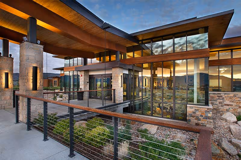 Modern senior living facility entrance with large glass windows, stone columns, wood overhangs, and a metal railing overlooking landscaped rocks at sunset.