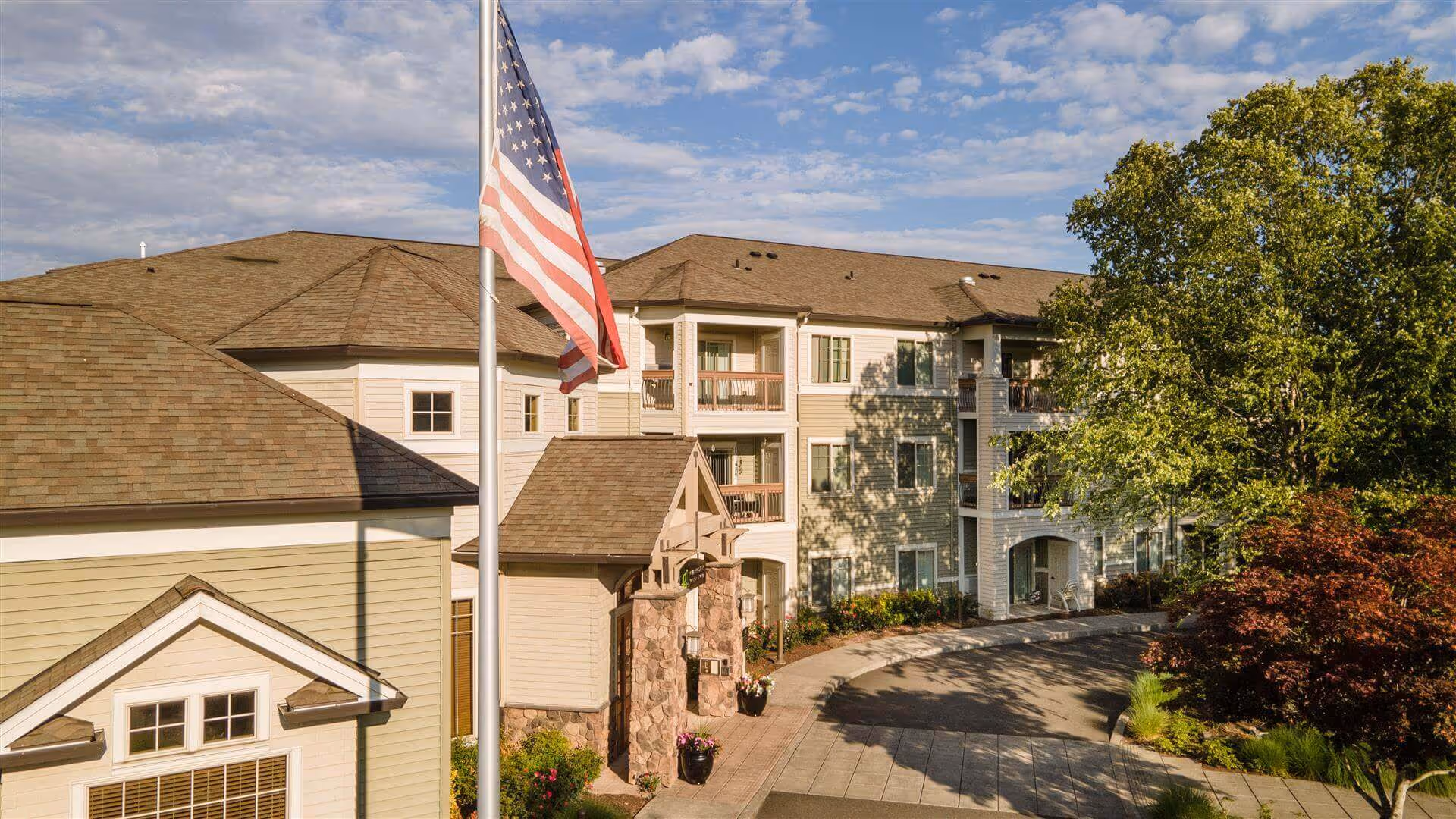 Exterior view of a multi-story senior living facility building with beige siding and stone accents, surrounded by trees and landscaping. An American flag is prominently displayed on a flagpole in front of the building under a partly cloudy sky.
