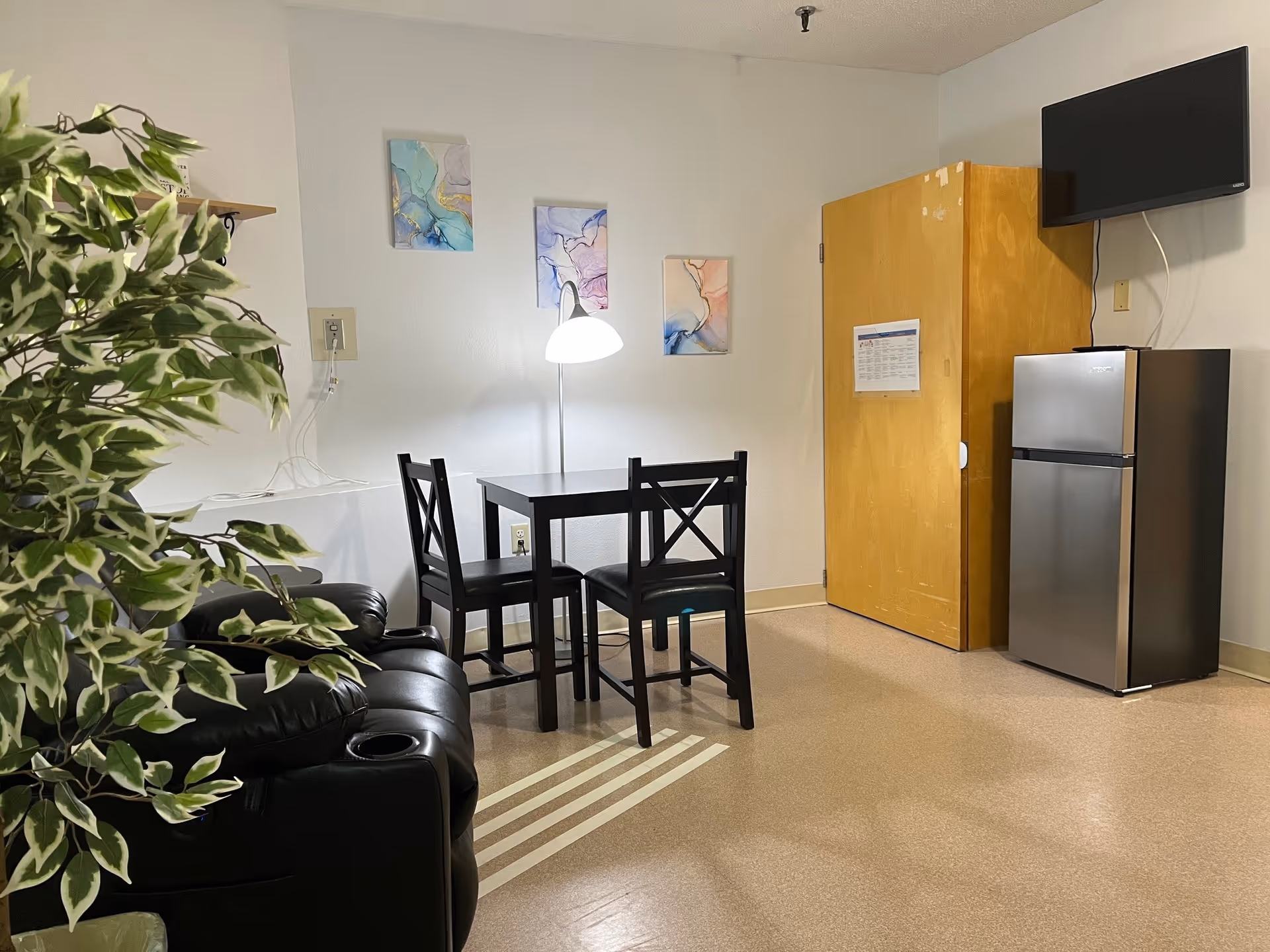 Interior of a room with a small black dining table and two chairs, a floor lamp, a black leather recliner chair, a small refrigerator, a wooden cabinet, a wall-mounted TV, and three abstract paintings on the wall. There is also a leafy green plant in the foreground.