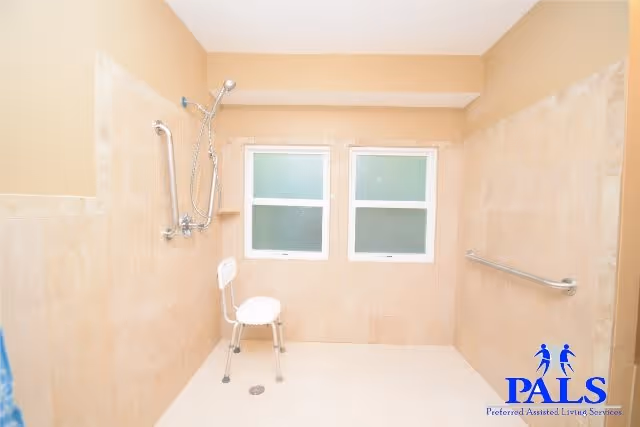 Accessible tiled shower room with grab bars, a shower chair, handheld shower head, and two frosted windows.