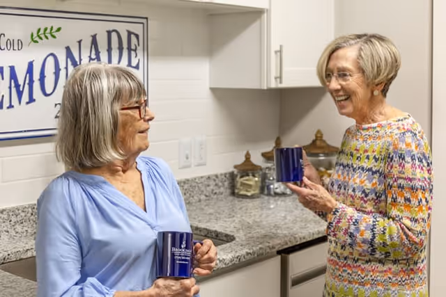 Two elderly women standing in a kitchen area, smiling and holding blue mugs. One woman is wearing a light blue blouse and glasses, and the other is wearing a colorful patterned sweater. Behind them is a granite countertop with jars and a sign on the wall that reads 'Cold Lemonade'.