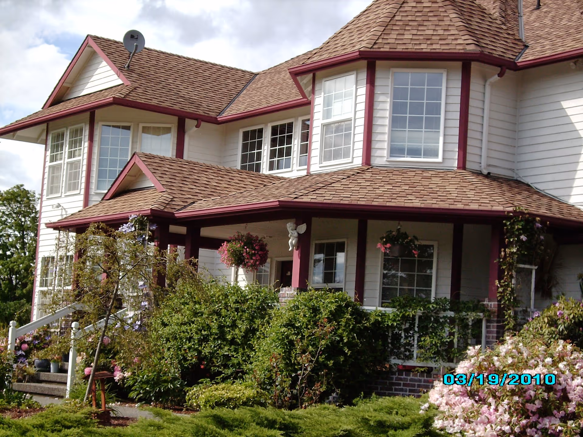 Front exterior of a two-story house with a covered porch, bay windows, and landscaped shrubs and flowers.