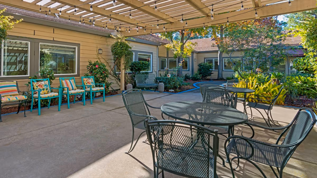 Outdoor patio area at Brookdale Roseville with metal tables and chairs under a pergola, surrounded by plants and trees, and a row of colorful cushioned chairs against the building wall.