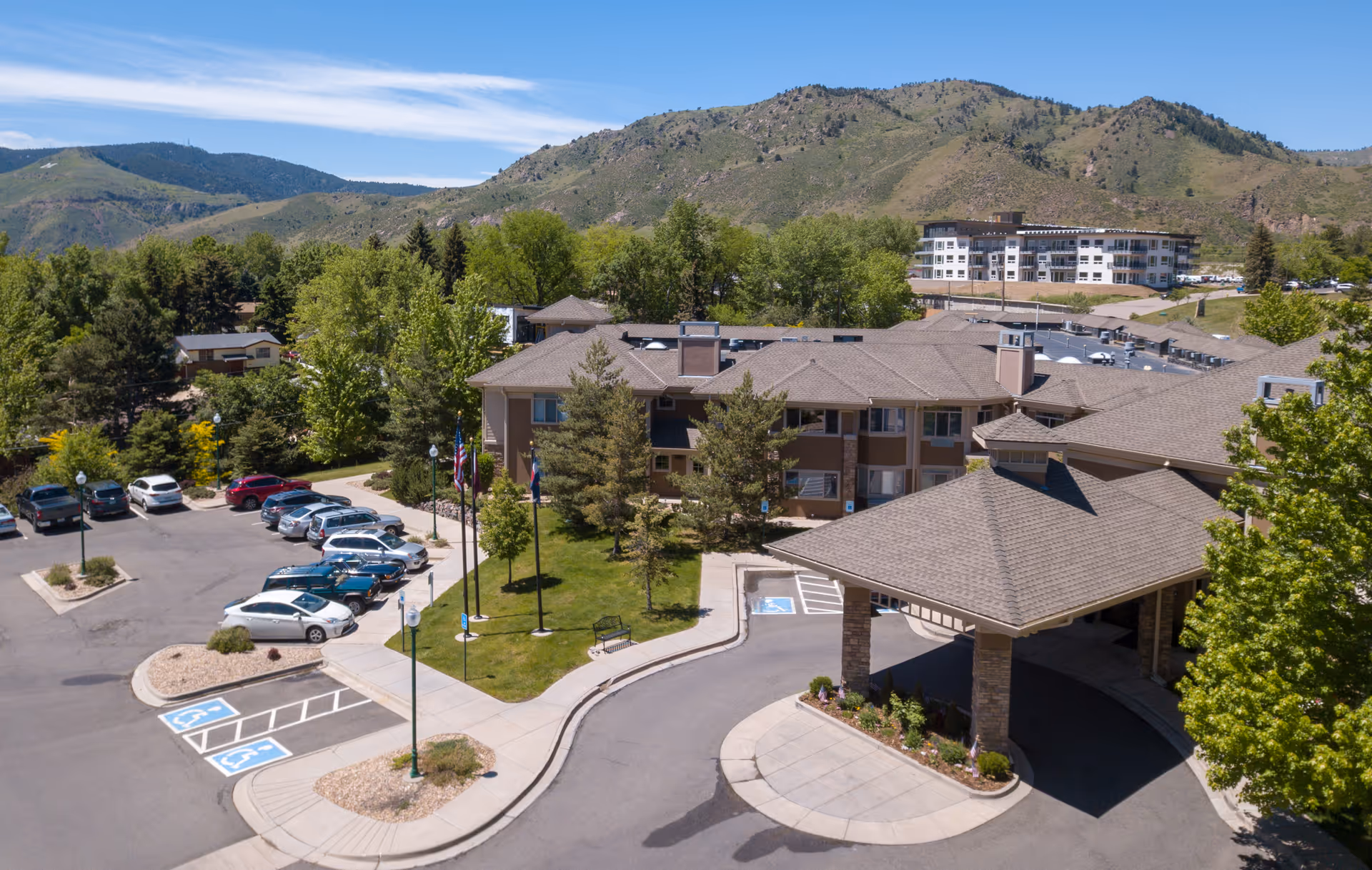 Aerial view of the senior living facility showing the main building with a covered entrance, parking lot, flags, and surrounding hills.