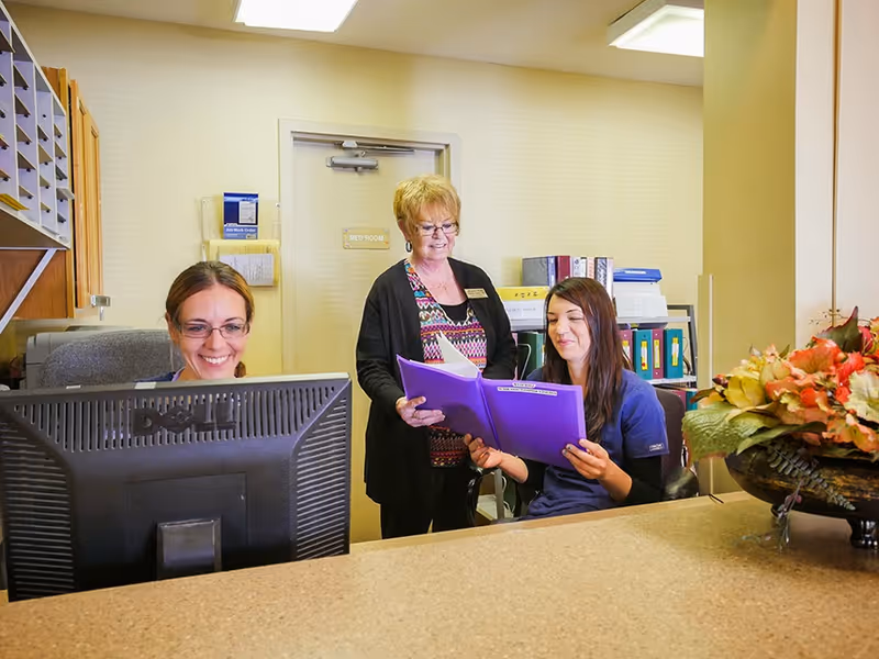 Three women at a reception desk in an office setting. One woman is seated at a computer smiling, another woman is standing and looking at a purple folder held by the third woman who is seated. There are shelves with files and a door labeled 'Med Room' in the background. A decorative flower arrangement is on the counter.