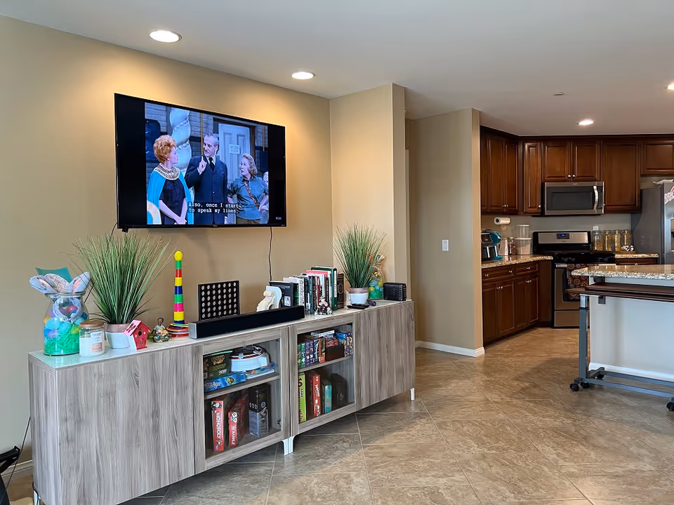 A cozy living area in a senior living facility featuring a wall-mounted flat-screen TV displaying a black-and-white show with subtitles. Below the TV is a light wood cabinet with glass doors containing board games and books. The cabinet top holds decorative items including two potted plants, a colorful stacking toy, a candle, and small figurines. To the right, there is a kitchen area with dark wood cabinets, stainless steel appliances, and granite countertops. The floor is tiled with beige tiles and recessed ceiling lights illuminate the space.