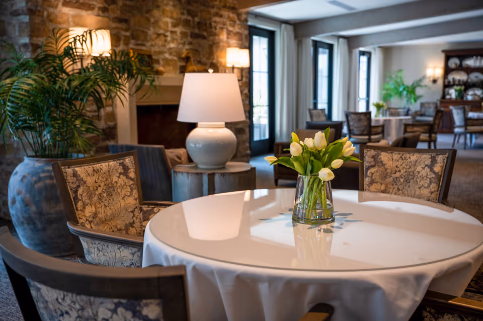 A cozy dining area with a round table covered with a white tablecloth and a vase of white tulips in the center. Surrounding the table are upholstered chairs with floral patterns. In the background, there is a stone fireplace, a large potted plant, a table lamp on a wooden side table, and more tables and chairs near large windows with curtains.