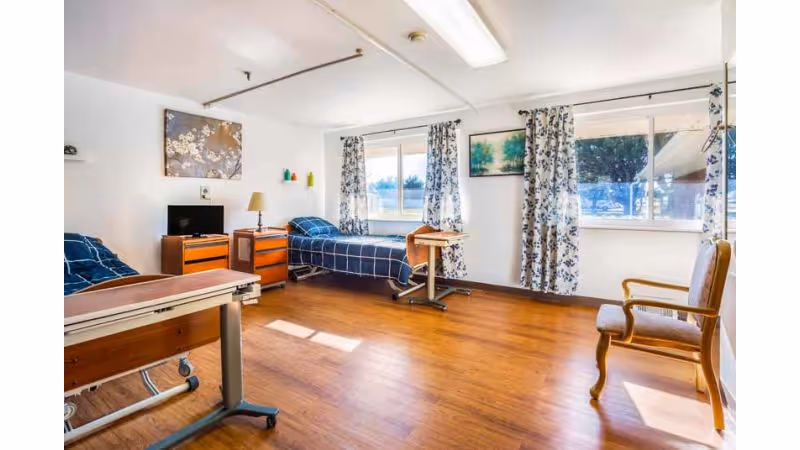A bright and spacious room with two hospital-style beds covered in blue checkered bedding, wooden floors, two windows with floral curtains letting in natural light, a wooden chair, a small TV on a wooden dresser, and a bedside table with a lamp.