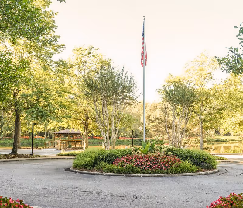 A landscaped roundabout with a flagpole flying the American flag in the center, surrounded by green bushes and pink flowers. In the background, there are trees, a wooden gazebo, and a small pond, all under bright daylight.