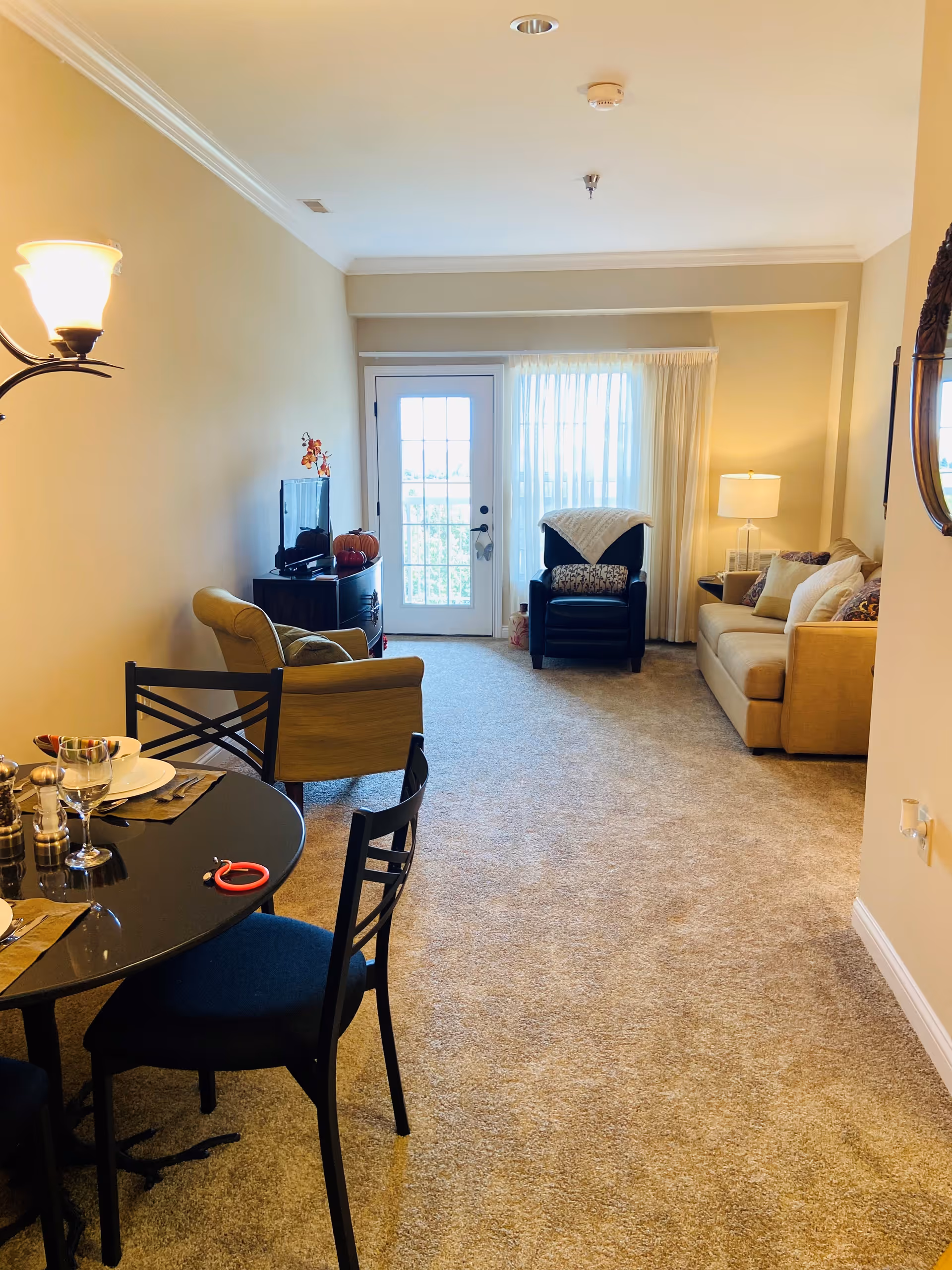 Bright carpeted living room with a sofa, armchairs, a TV stand, and a small dining table, centered on a glass door leading to a balcony.
